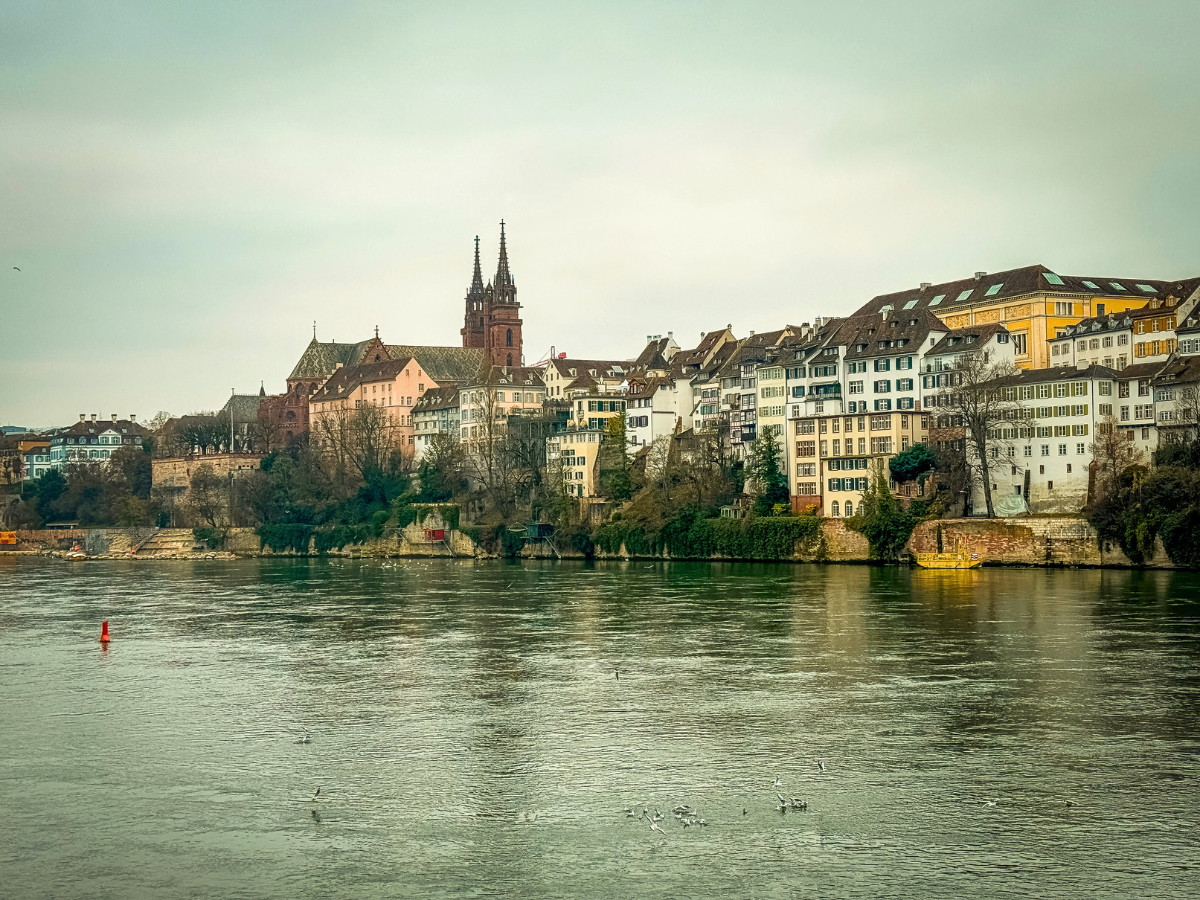 Colorful historic buildings and the red sandstone spires of Basel Minster line the Rhine River under a cloudy sky in Basel, Switzerland.