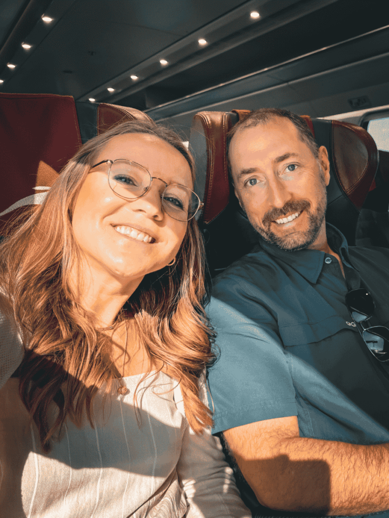 Kate and a man smile at the camera while seated in a modern European train with red leather headrests and soft overhead lighting.