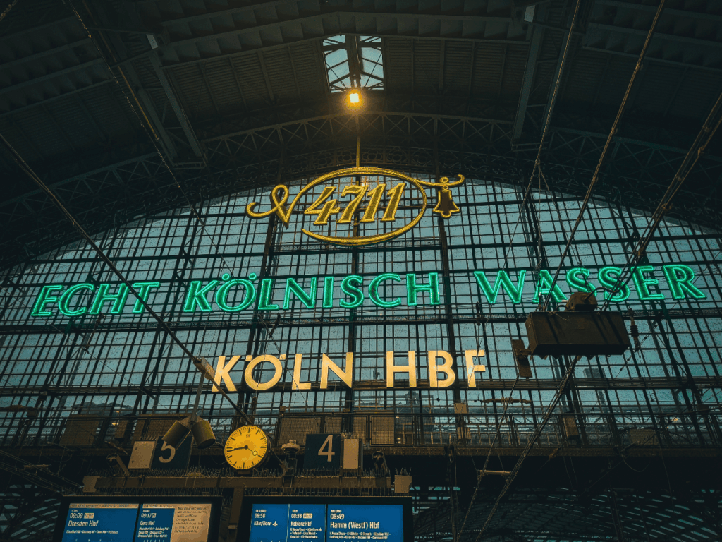 The illuminated signs inside Köln Hauptbahnhof (Cologne Central Station) display "4711 Echt Kölnisch Wasser" in green neon and "KÖLN HBF" in white letters against a tall glass wall above the train platforms.
