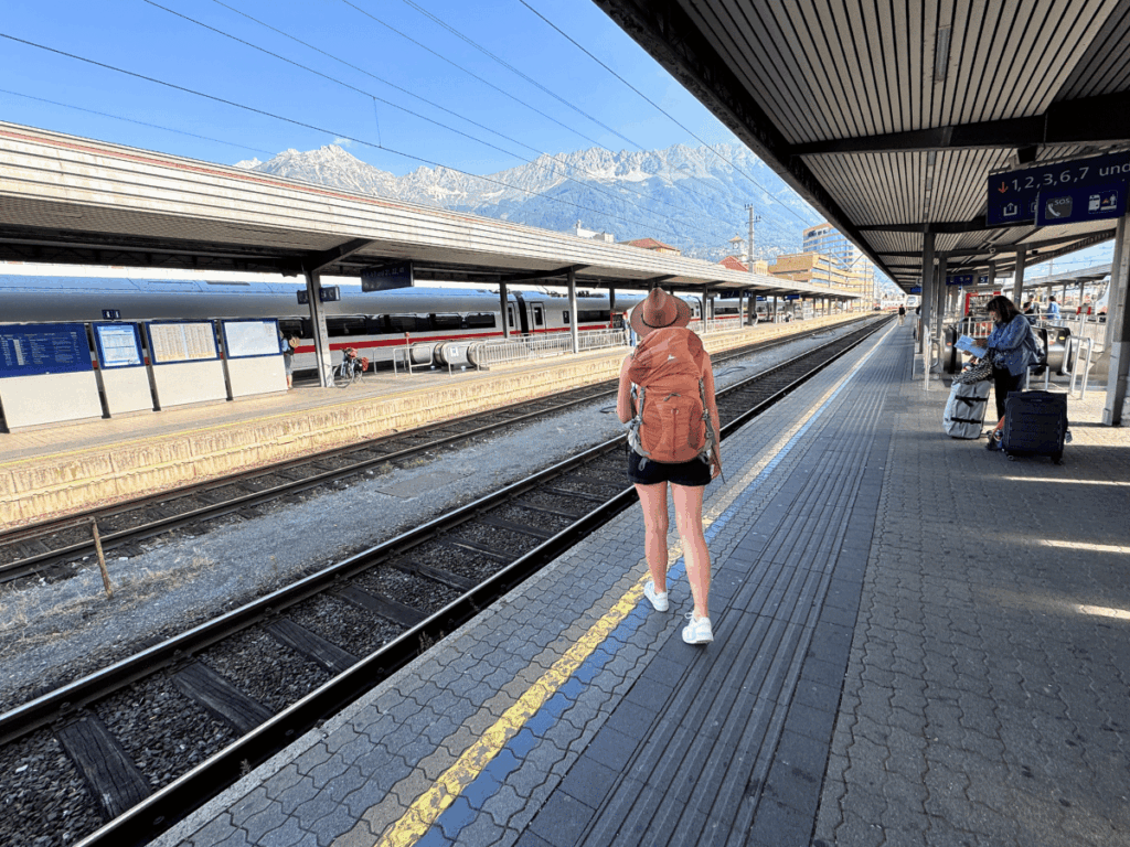 Kate walks along a European train platform with a large backpack and hat, as snow-capped mountains rise behind the station under a clear blue sky.