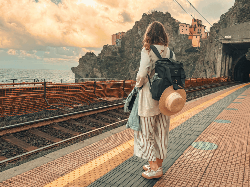 Kate stands at a colorful coastal train station in Cinque Terre, facing the sea and cliffside buildings with a backpack and sunhat on her back.