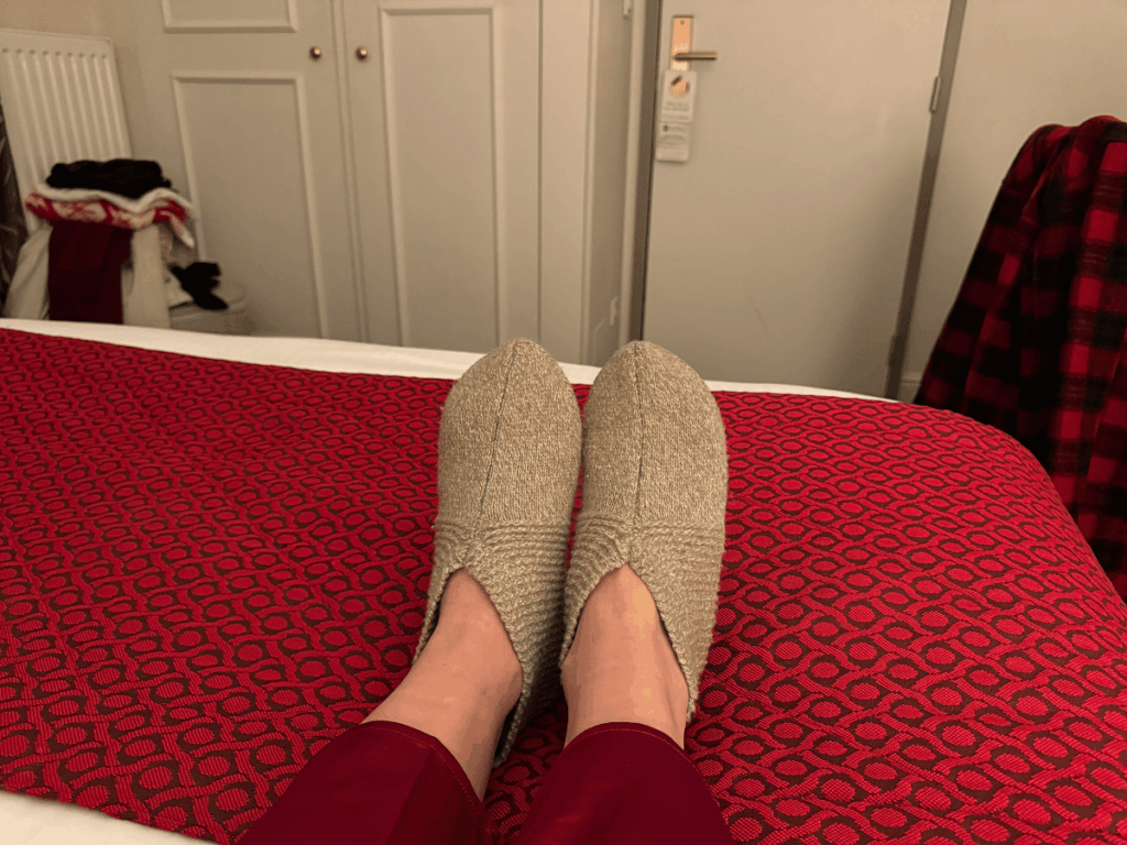 A person wearing cozy beige slippers and red pajama pants rests their feet on a hotel bed with a red patterned throw, capturing a moment of winter travel relaxation indoors.