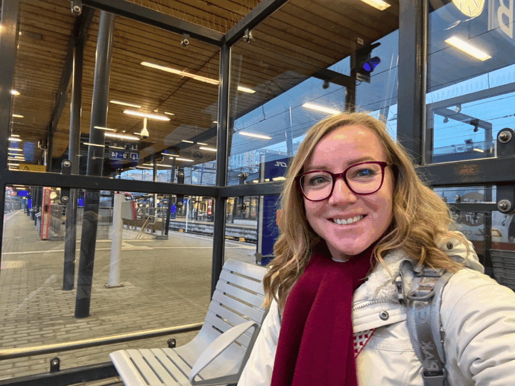 Kate smiles while sitting in a glass-enclosed waiting area at a European train station, wearing a white winter coat and red scarf with train platforms visible through the windows.