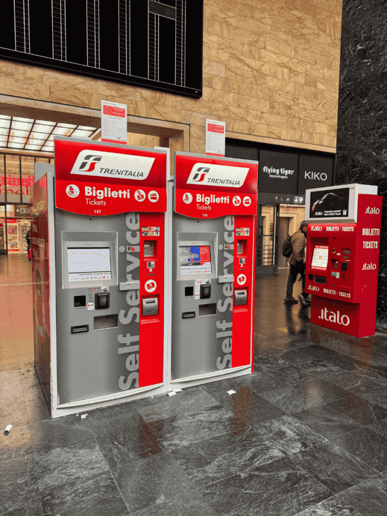 Three red self-service ticket machines for Trenitalia and Italo stand inside an Italian train station, with brand logos and multilingual instructions displayed above the screens.