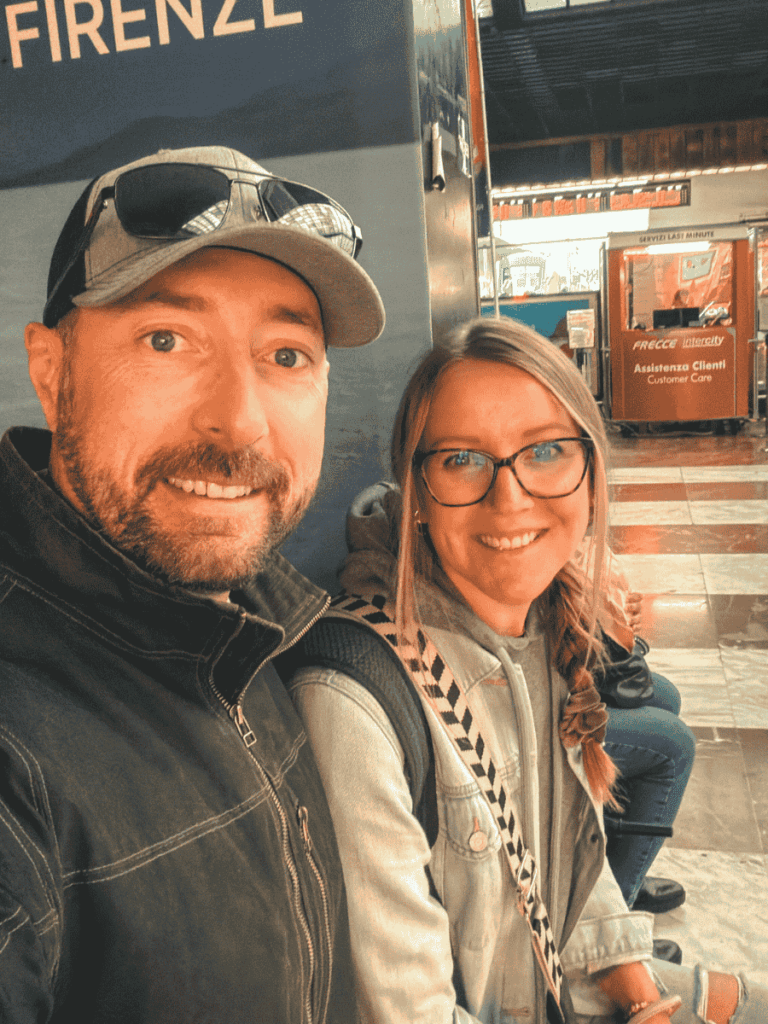 Kate and a man smile while seated inside Firenze Santa Maria Novella train station, with the “Customer Care” desk and signage in Italian visible behind them