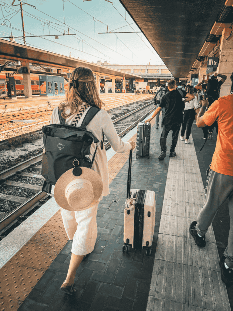 Kate walks along a busy European train platform pulling a suitcase, with a backpack and sunhat on her back, surrounded by other travelers waiting to board.