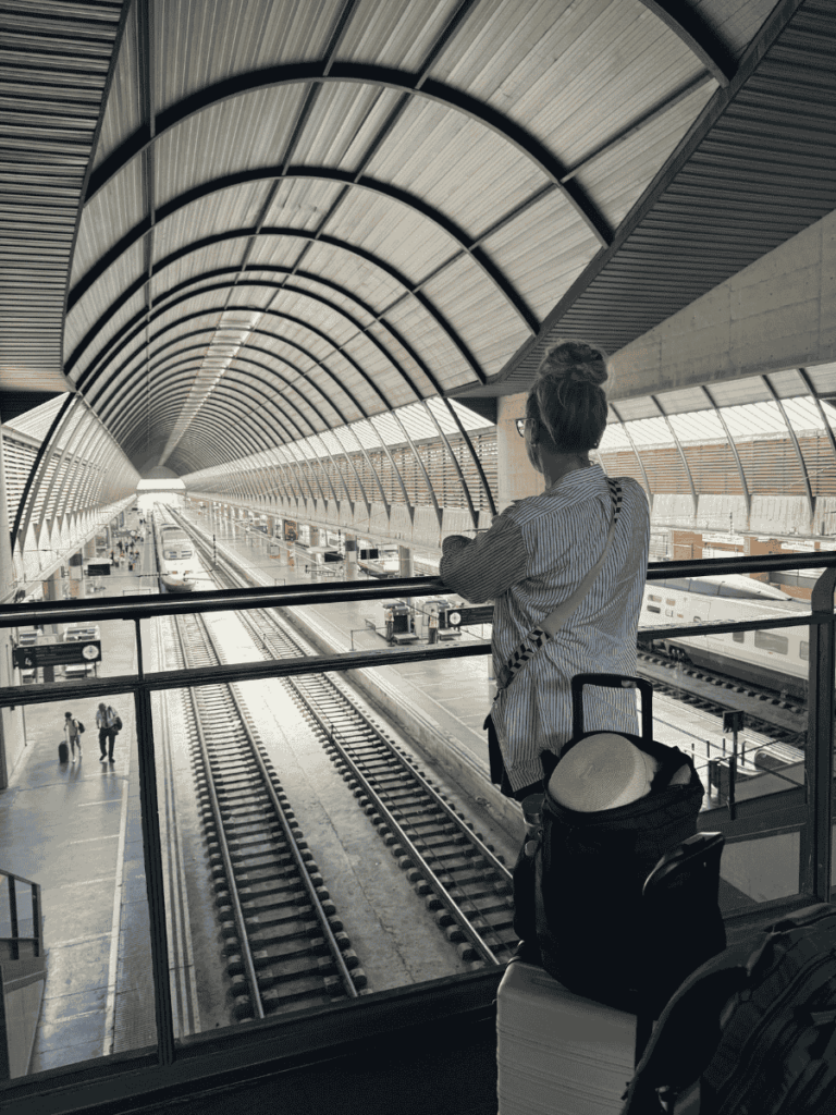 From an upper level, Kate looks out over a modern, arched train station as parallel tracks stretch into the distance, with her suitcase and backpack beside her.