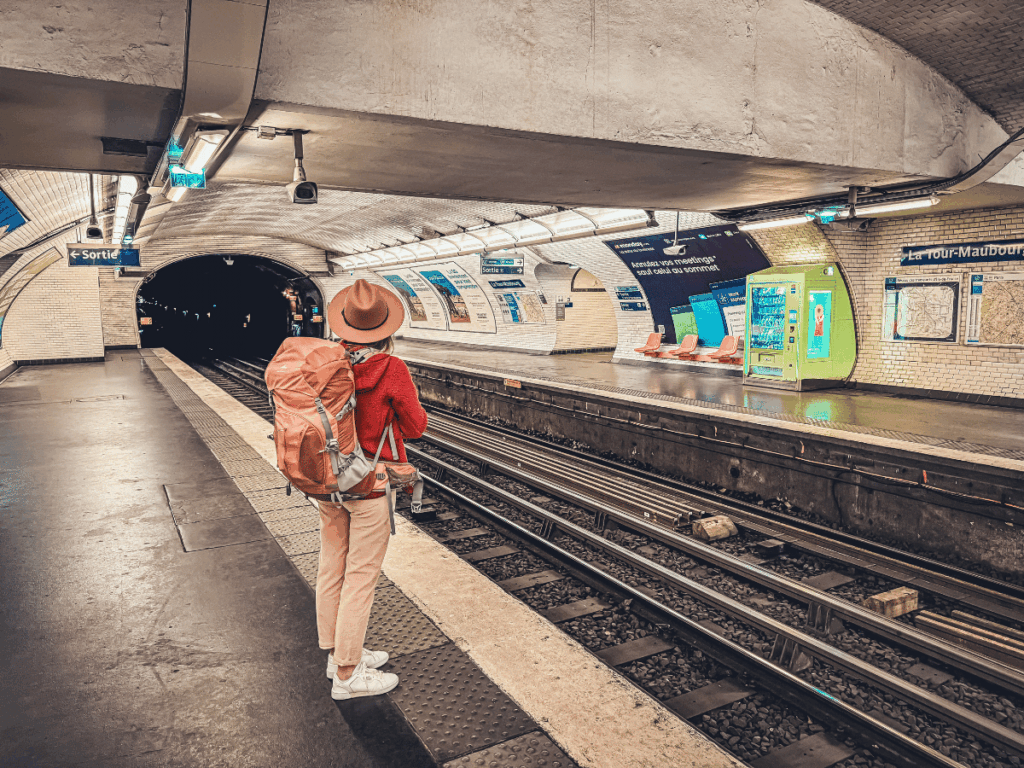 Kate stands on the platform of a Paris metro station with a large rust-orange backpack and wide-brim hat, facing the dark tunnel at La Tour-Maubourg stop under arched tiled ceilings.