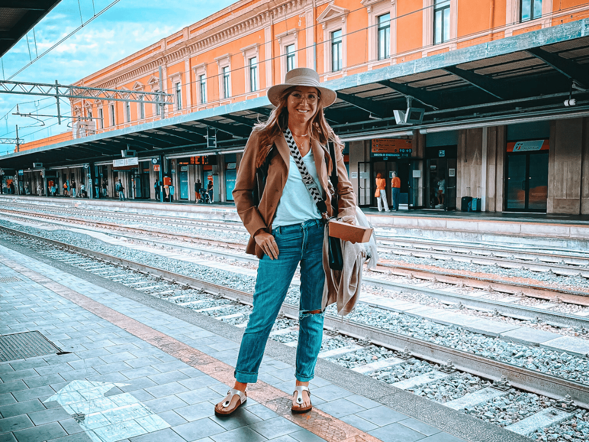 Kate stands on a European train platform in casual travel attire, including a wide-brim hat and crossbody bag, with train tracks and a historic orange station building in the background.