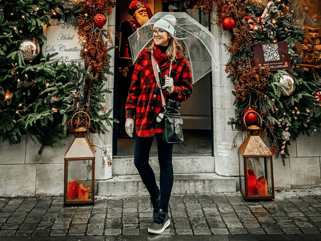 Kate is dressed in a red plaid jacket, mint beanie, gloves, and winter boots holds a clear umbrella while standing in front of a festively decorated hotel entrance with Christmas garlands and lanterns.