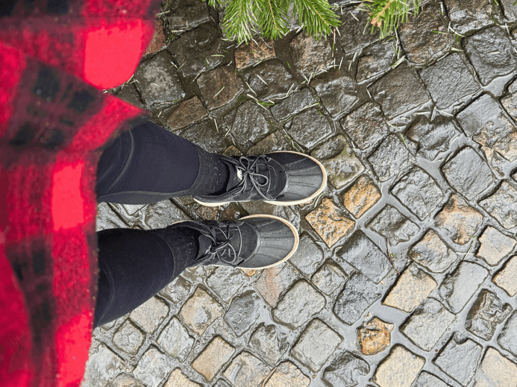 Looking down at waterproof black duck boots on wet cobblestone, the image shows a winter travel outfit with black leggings and a red plaid coat, perfect for staying dry on rainy streets.
