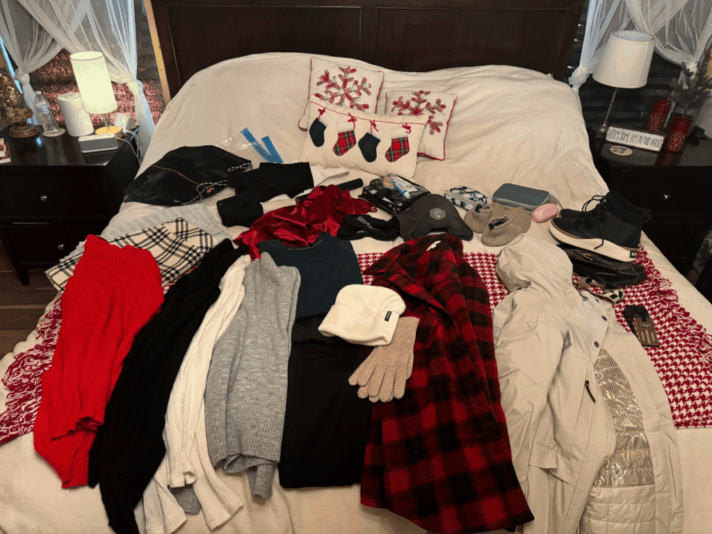 A neatly laid-out winter travel wardrobe on a bed includes sweaters, scarves, gloves, thermal layers, a white winter coat, waterproof boots, toiletries, and accessories, all arranged beside festive holiday pillows.