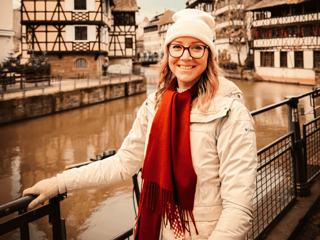 Kate in a white winter coat, red scarf, white beanie, and gloves smiles by a riverside railing, with half-timbered houses in the background on a cold day in Europe.