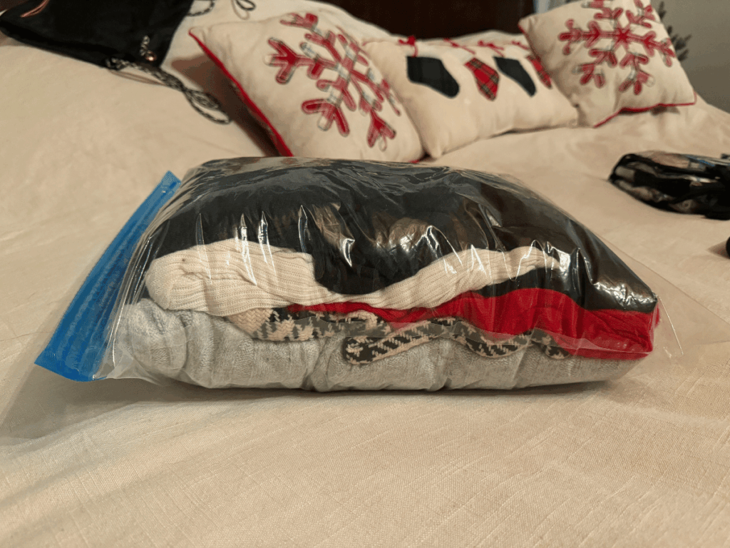 A stack of winter clothes, including sweaters and thermals, is tightly packed into a clear compression bag on a bed with festive snowflake pillows in the background.