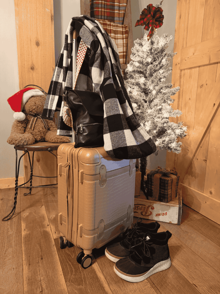 A beige rolling suitcase is topped with a black leather backpack and a black-and-white plaid scarf, set beside winter boots, a frosted white Christmas tree, and cozy holiday decor, capturing winter travel essentials.