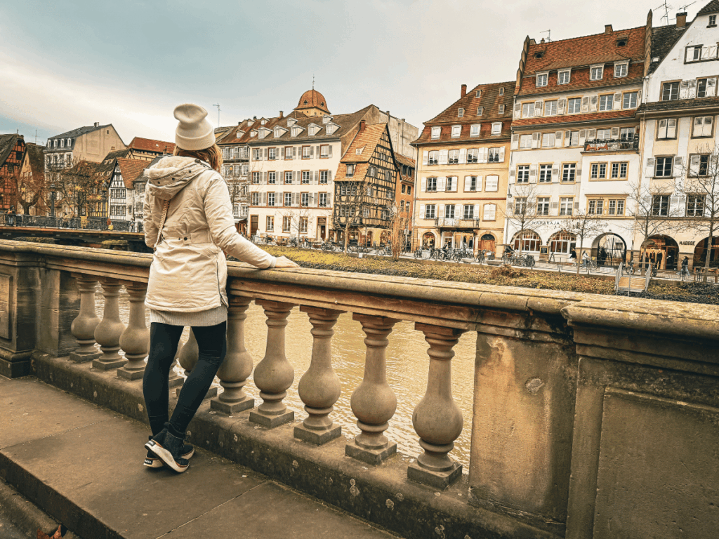 A woman dressed in a white winter coat, black leggings, and beanie stands at a stone railing overlooking a river and a row of colorful European buildings on a cloudy winter day.