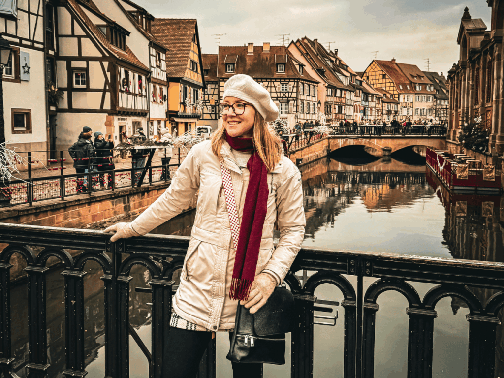 A woman in a winter coat, red scarf, gloves, and beret smiles while leaning on a black railing over a canal, with half-timbered buildings and a festive crowd in the background.