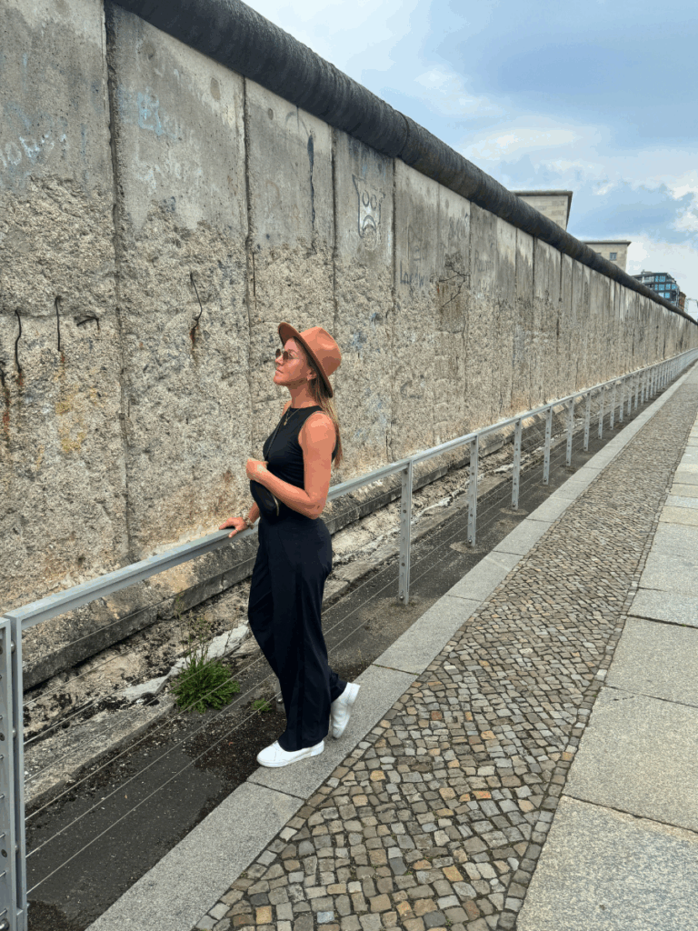 Kate walking alongside the Berlin Wall wearing high-waisted black travel pants with a relaxed fit, paired with white sneakers and a tan hat.