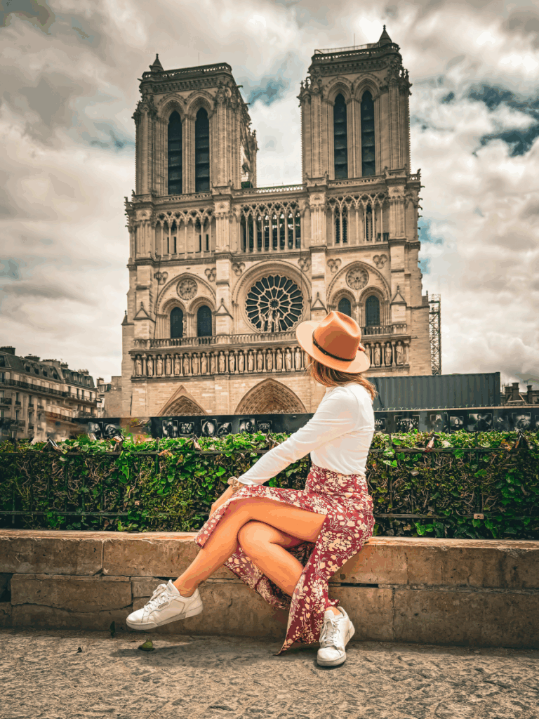 Kate sitting in front of Notre-Dame Cathedral wearing stylish white leather sneakers with light gold detailing, paired with a floral skirt and a tan hat.