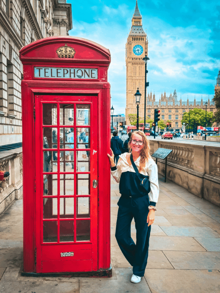 Kate is posing by a red London phone booth, wearing navy drawstring travel pants with a relaxed fit, styled with white sneakers and a color-blocked jacket.