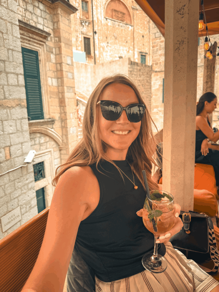Kate smiling at an outdoor café while wearing a fitted black sleeveless tank top, paired with striped pants and oversized sunglasses.