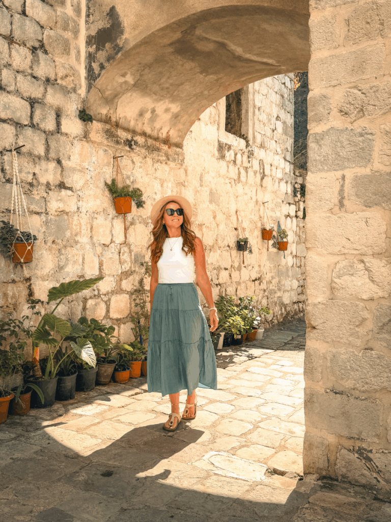 Kate walking through a sunlit stone alleyway wearing a fitted white sleeveless tank top, paired with a flowy teal skirt, sandals, and a straw hat.