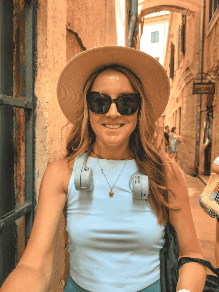 Kate exploring a narrow alleyway wearing a beige sun hat and a compact gray neck fan with dual vents for portable, hands-free cooling.