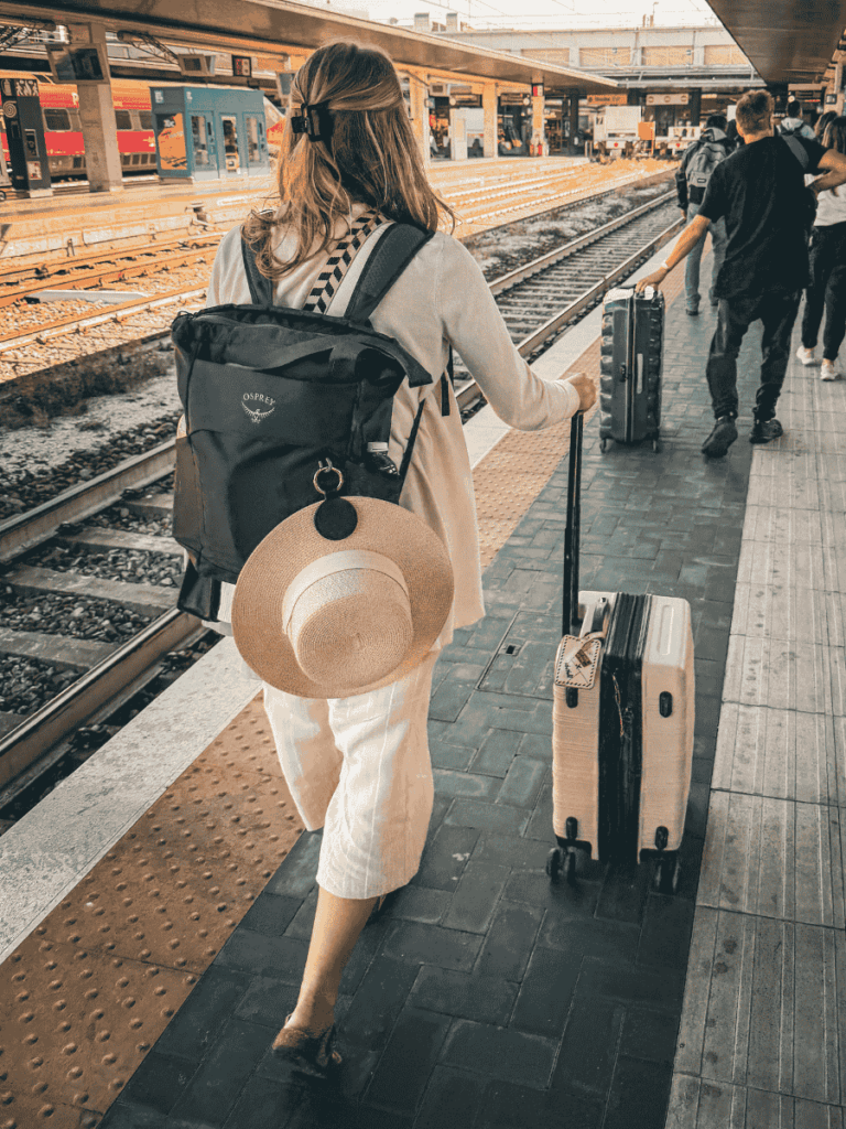 Kate walking along a train platform with a straw hat securely fastened to a black Osprey backpack using a sleek black hat clip.