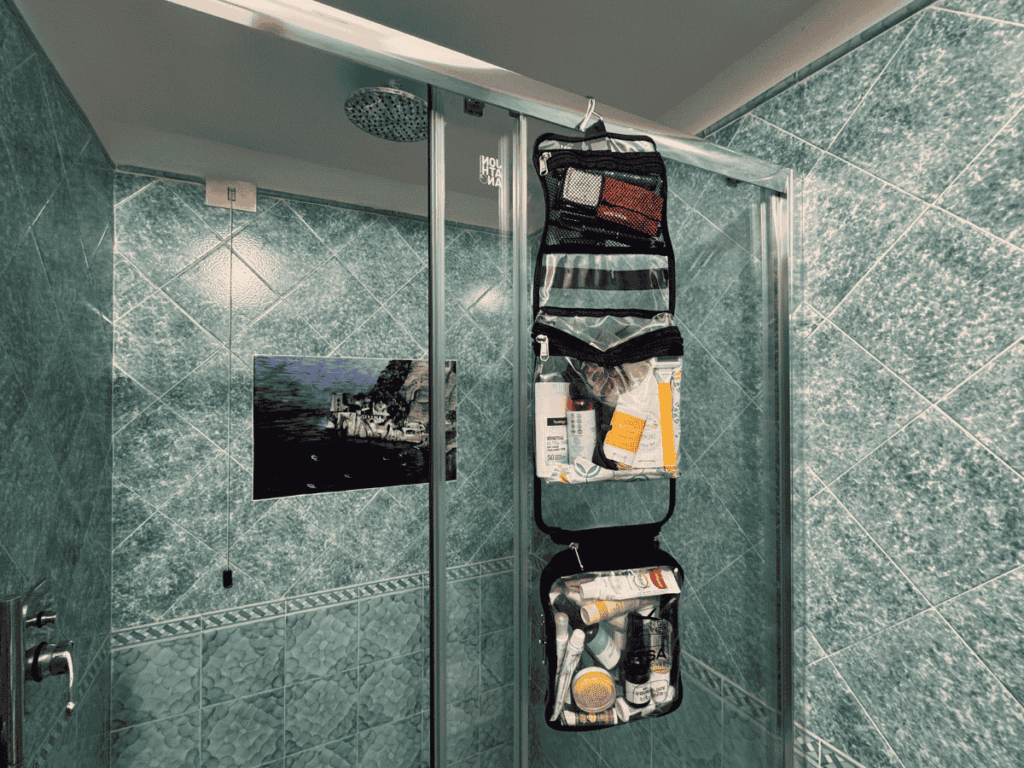 Hanging travel toiletry bag fully stocked with personal care essentials, neatly organized in clear zippered compartments and hooked on a glass shower door for easy access.