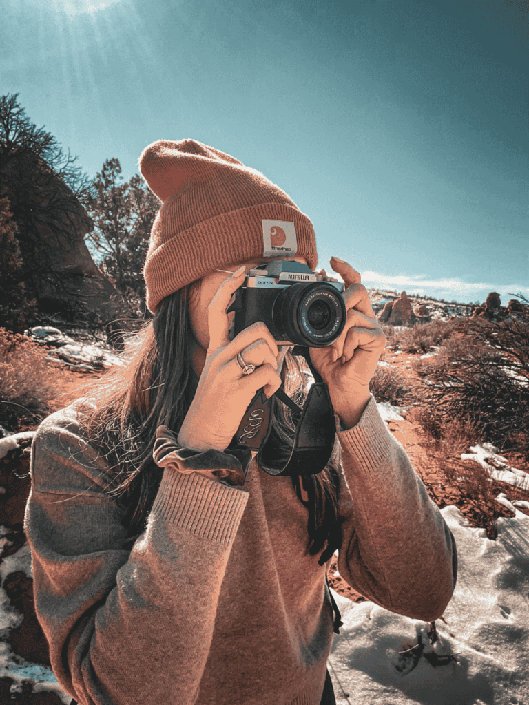 Kate is outdoors in a beanie and sweater, holding a Fujifilm camera up to her face, ready to capture a shot in a rugged, snowy landscape.