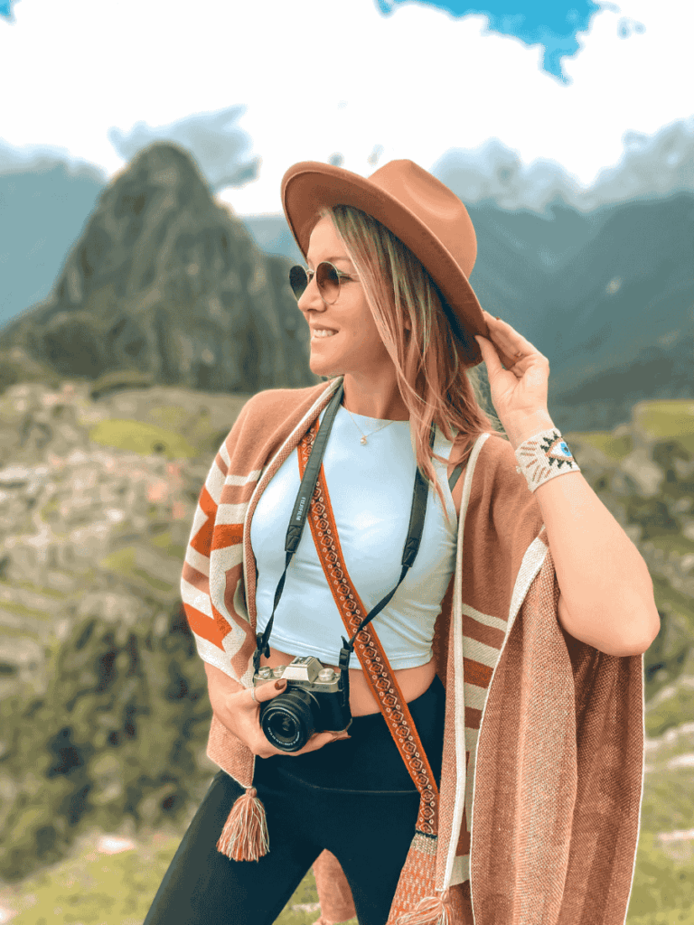 Kate posing with a Fujifilm camera hanging from her neck by a woven strap, smiling against a mountain backdrop while adjusting her wide-brim hat.