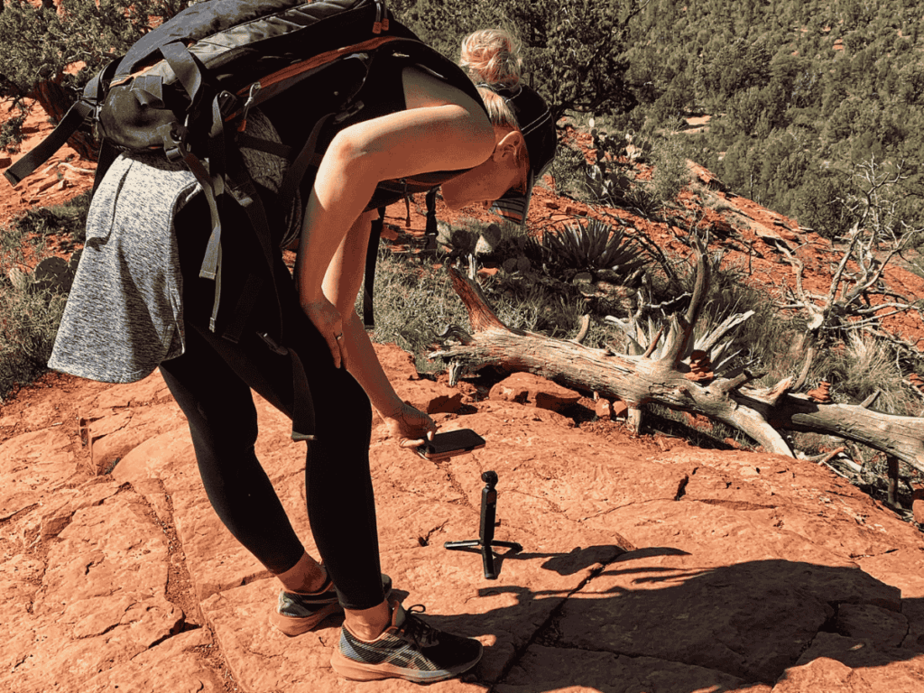 Kate is on a rocky hiking trail using a compact action camera mounted on a mini tripod to capture footage, with a large backpack and scenic desert landscape in the background.