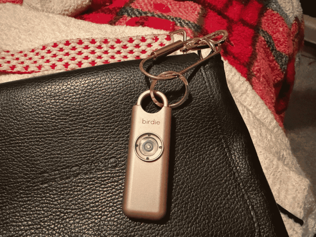 Close-up of a rose gold Birdie personal safety alarm clipped to a black leather bag with a carabiner, resting on a patterned red and white blanket.