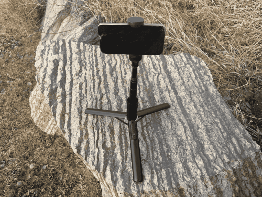 Compact black tripod with a smartphone mounted on top, set up on a textured stone surface in a dry grassy outdoor setting, ready for stable photo or video capture.