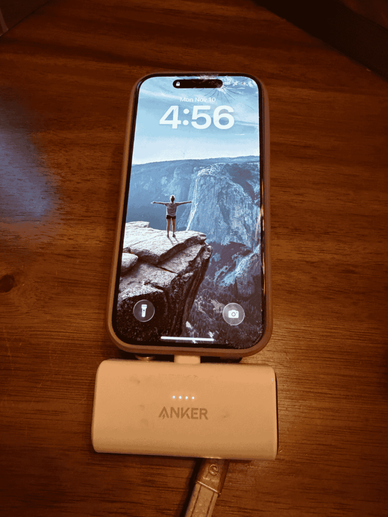 iPhone plugged into a compact white Anker Nano power bank charger, with LED indicators lit up, resting on a wooden surface.