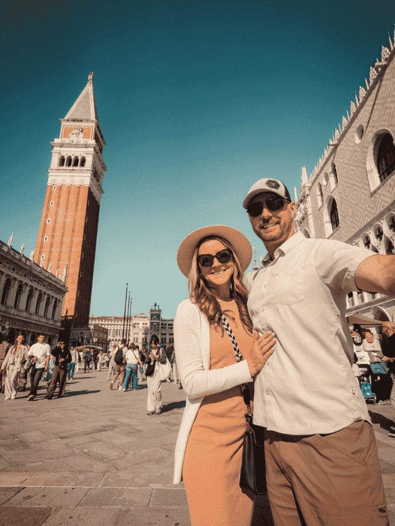 Kate wearing a white cardigan over a peach dress, standing beside a man in front of St. Mark's Campanile in Venice.