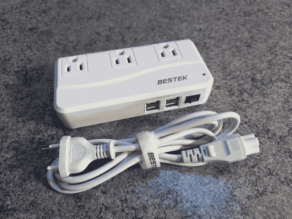 White BESTEK travel power adapter featuring three AC outlets and four USB ports, shown with a neatly coiled detachable power cord on a dark textured surface.
