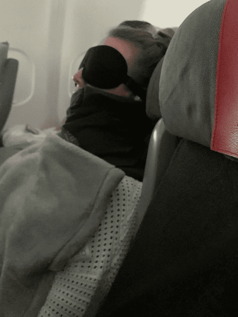 Kate sleeping on a plane using the Trtl travel pillow for neck support, paired with an eye mask and blanket for comfort.