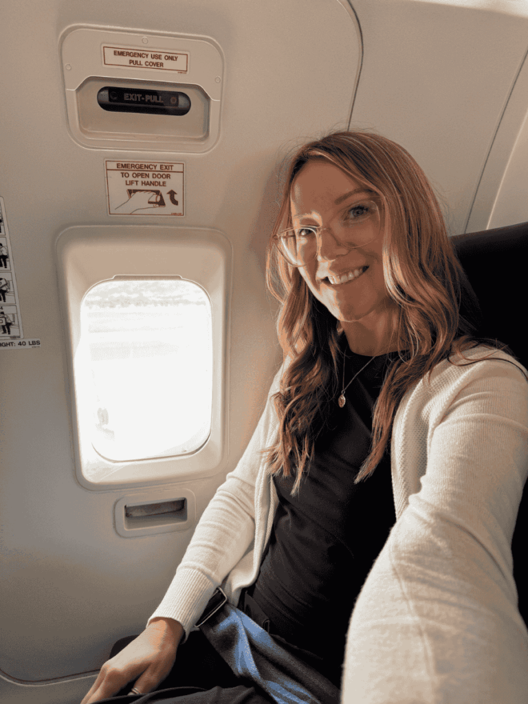 Kate sitting by the airplane window wearing a white cardigan and black outfit, smiling during a daytime flight.