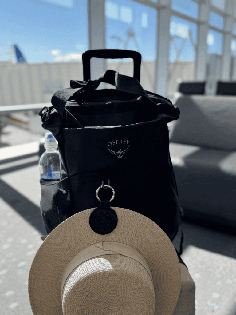 Black Osprey backpack resting on top of a rolling suitcase at an airport gate, with a straw hat clipped to the front and a water bottle in the side pocket.