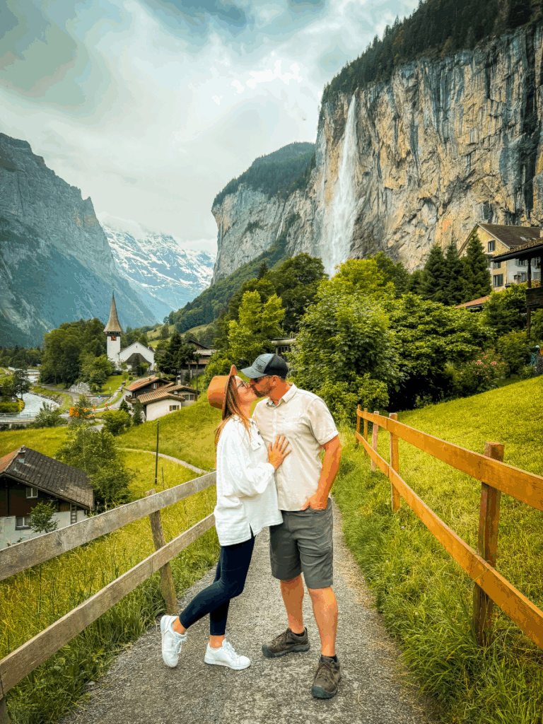Kate wearing crisp white sneakers with subtle gold details, standing on a mountain path while kissing her partner with a scenic waterfall and alpine village in the background.
