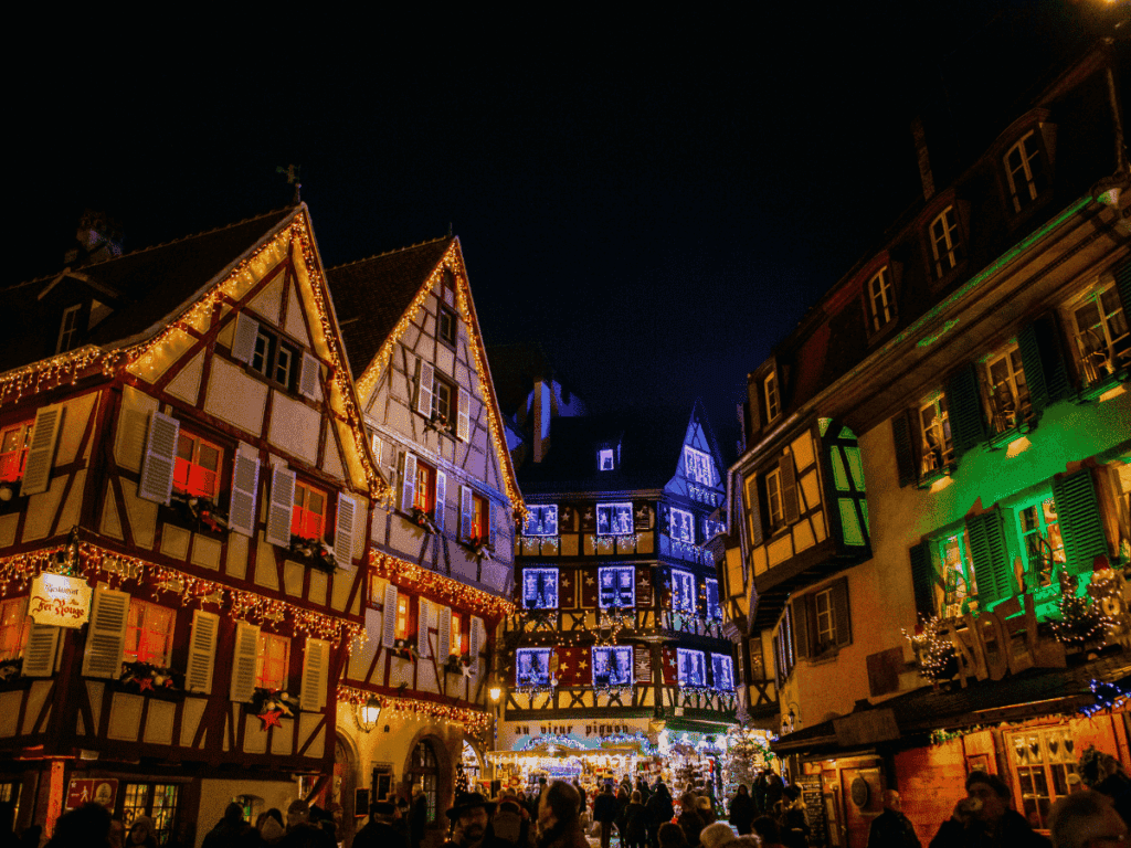 Half-timbered buildings in Colmar glow with colorful Christmas lights and window displays at night.