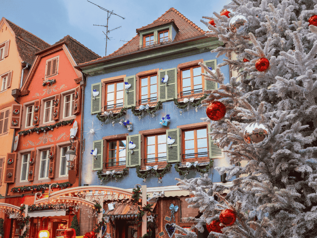 Colorful, festive buildings in Colmar are decorated for Christmas with garlands, wreaths, and a large frosted tree covered in red and silver ornaments.