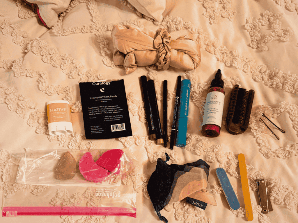 A flat lay of travel toiletries and beauty items, including makeup, dry shampoo, deodorant, hair tools, and a spot patch kit, spread out on a textured cream bedspread.