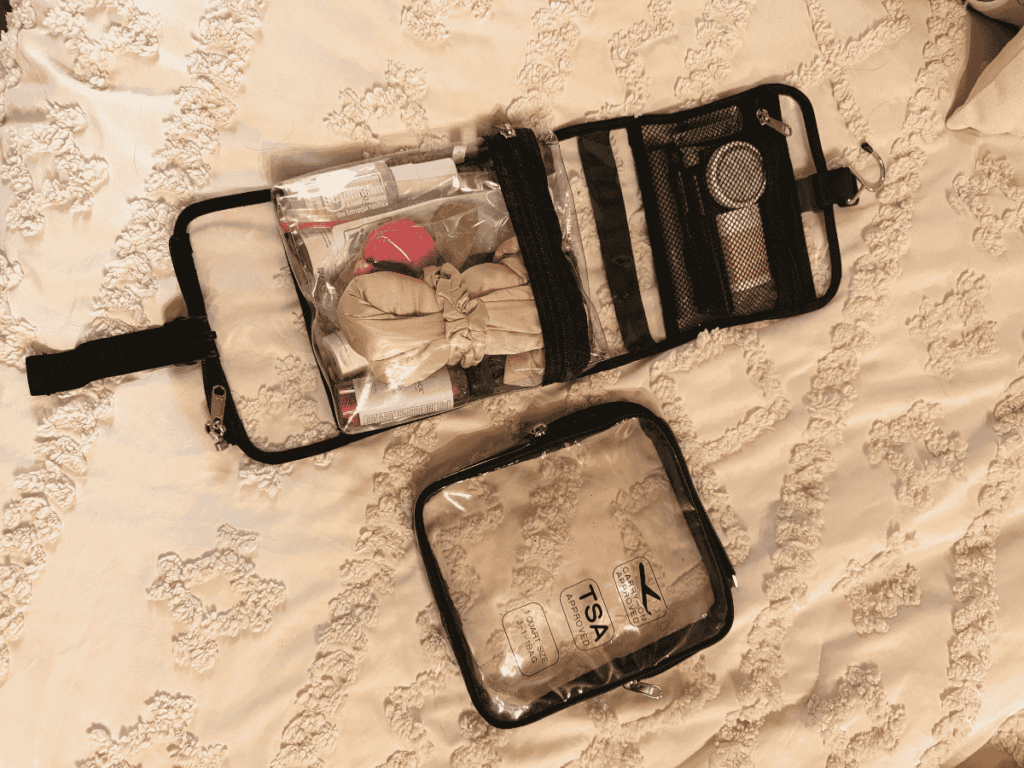 A fully packed hanging toiletry bag and a clear TSA-approved travel pouch are laid out on a cream-colored textured bedspread.