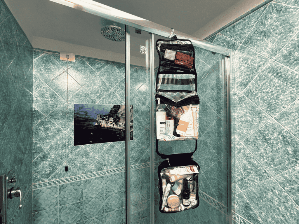 A hanging toiletry bag filled with travel-size essentials is hooked onto a glass shower door in a bathroom with teal tile walls.
