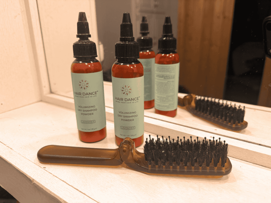 Three bottles of Hair Dance Volumizing Dry Shampoo Powder in lavender scent sit on a vanity beside a brown bristle hairbrush, reflected in a mirror.