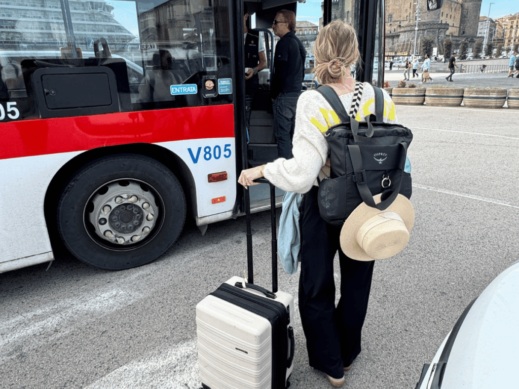 Kate stands with her suitcase and backpack, boarding a red and white city bus near a European port.