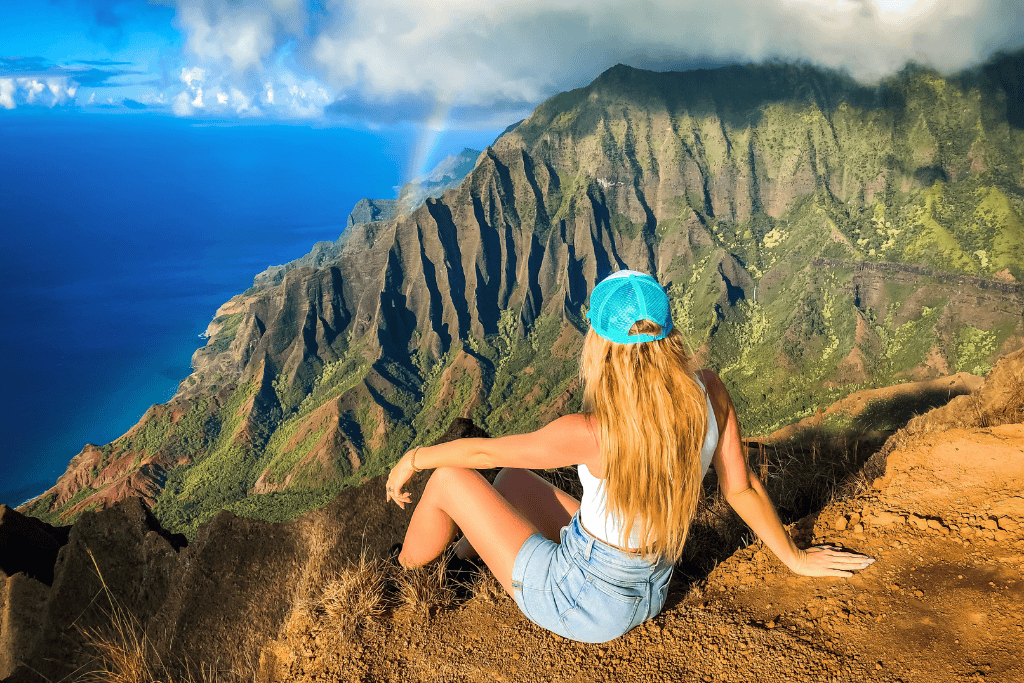 Kate, in a blue cap and denim shorts, sits at a breathtaking lookout, gazing at the towering emerald cliffs and deep valleys of the Nā Pali Coast with a faint rainbow in the misty sky.