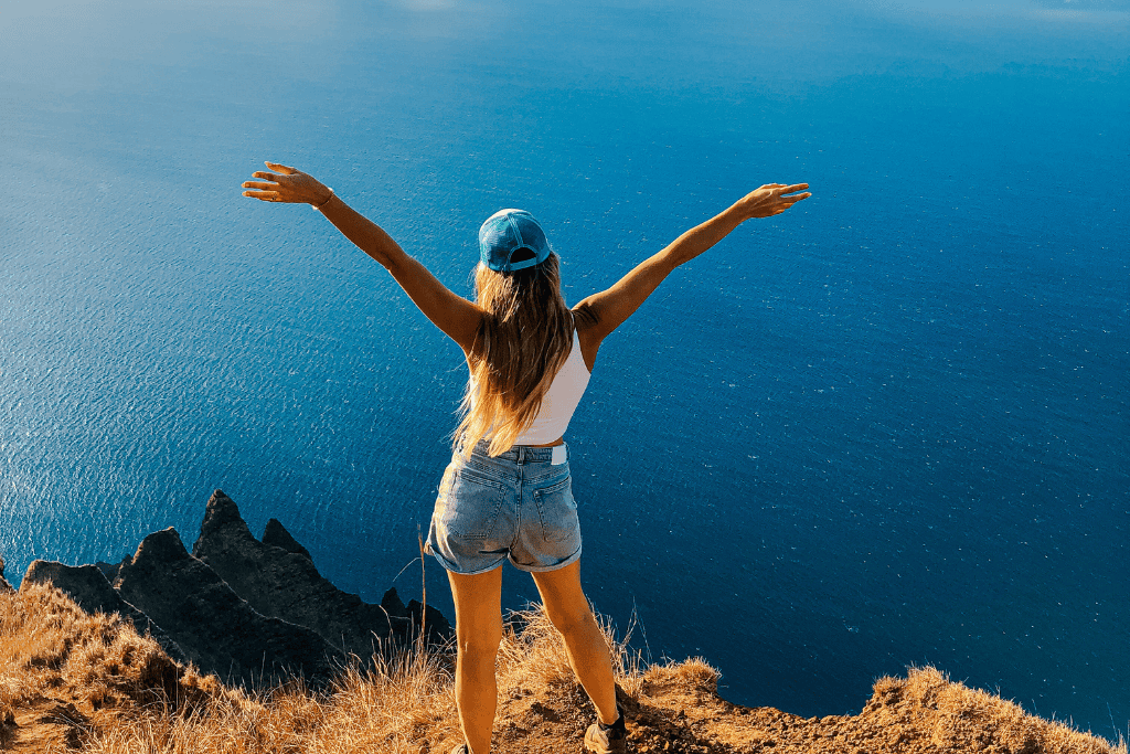 With arms stretched wide, Kate stands triumphantly on a cliff edge above the vast, calm Pacific Ocean, taking in the sheer magnitude of the Nā Pali Coastline's rugged peaks and endless blue horizon.